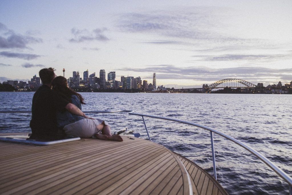 Cruise boat Sydney: Couple sitting close together on the teak bow of a boat, watching a city skyline and harbour bridge