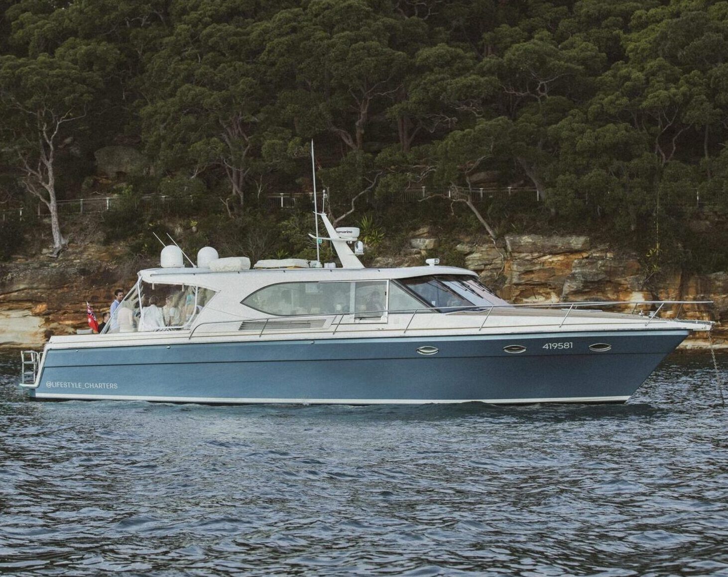 Blue and white motor yacht Felix moored on calm water beside a bushy sandstone shoreline.