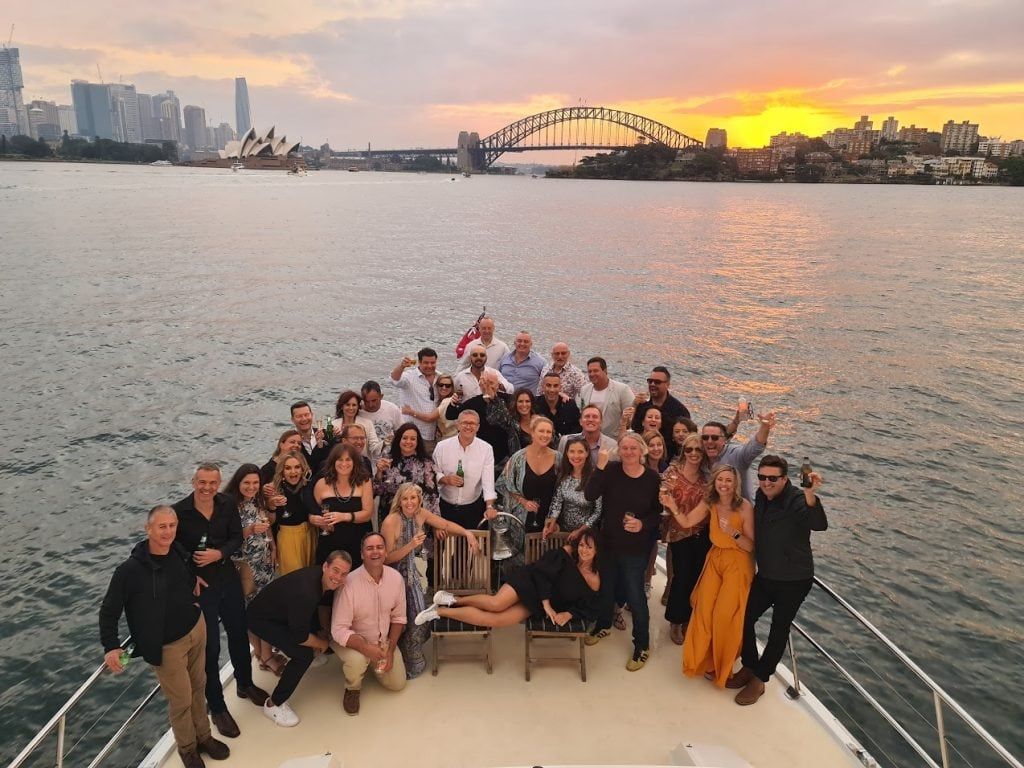 Large group posing with drinks on a boat's bow deck at sunset, with a harbour bridge and city skyline behind.