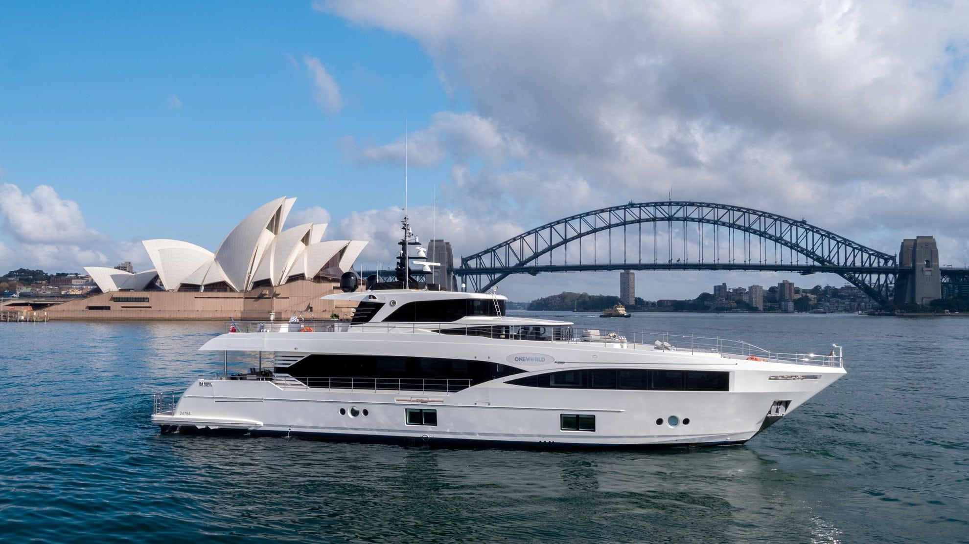 White superyacht Oneworld cruising on harbour water with the Sydney Opera House and Harbour Bridge behind.