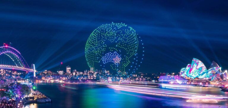 Drone light show forming a green and blue globe above a harbour at night, with lit bridge and illuminated opera house