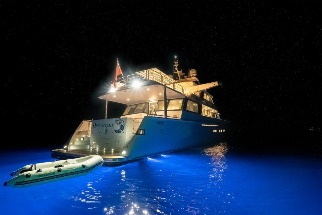 Motor yacht anchored at night with underwater lights turning the surrounding water bright blue, tender alongside.