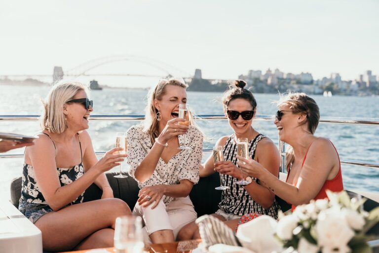Four women laughing with champagne glasses on the rear deck of a boat, harbour bridge and city skyline behind.