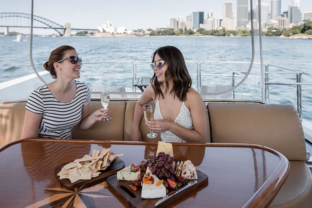 Eastern pontoon circular quay: Two women with sunglasses hold sparkling wine on a boat's rear lounge, harbour bridge and
