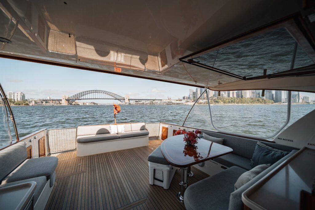 Hiring a boat sydney: Covered aft deck of a boat with teak flooring, lounge seating and a table, looking out to a harbour