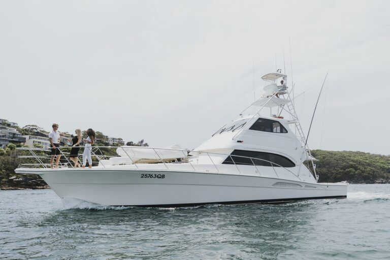 Four people stand on the bow of a white flybridge motor yacht cruising past a wooded shoreline.