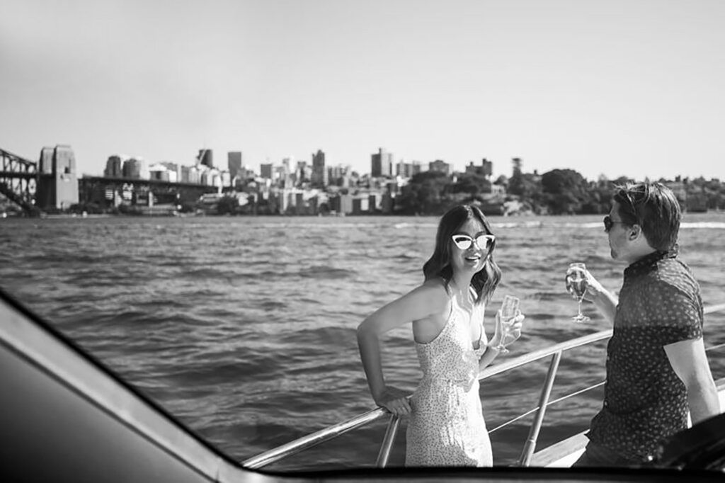 Rose bay wharf: Woman and man in sunglasses holding sparkling wine glasses on a boat deck with a city skyline behind