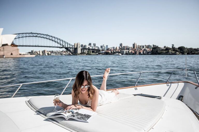 Woman in sunglasses lying on a boat's white bow cushion with a champagne glass and open magazine, harbour bridge behind.