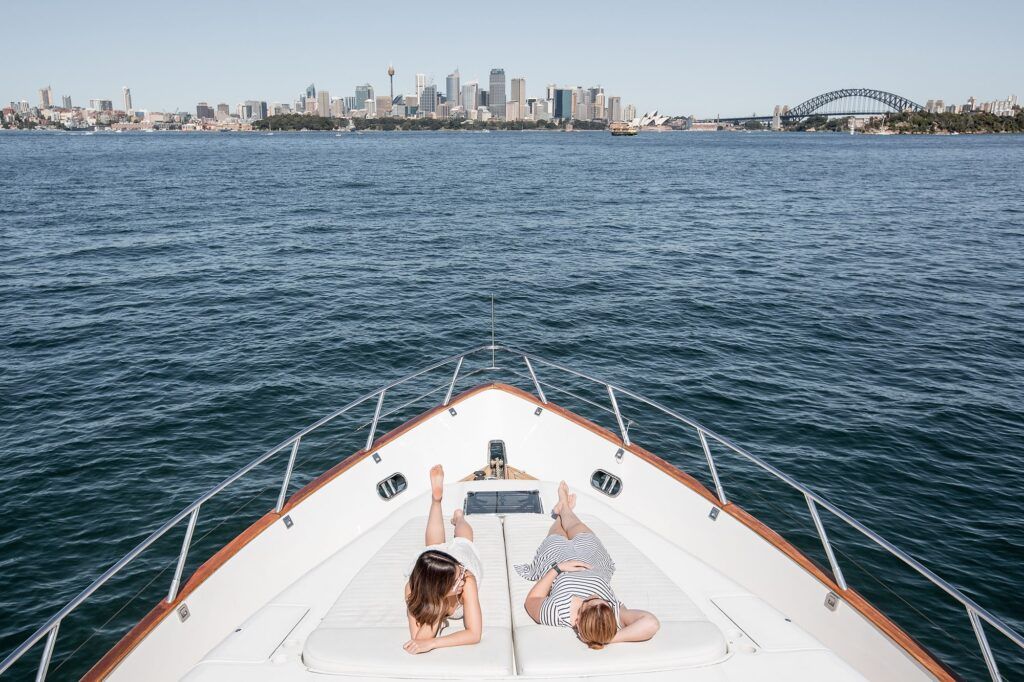 Winter boat party sydney: Two people lie facing forward on the bow sun pad of a white motor yacht, with a city skyline