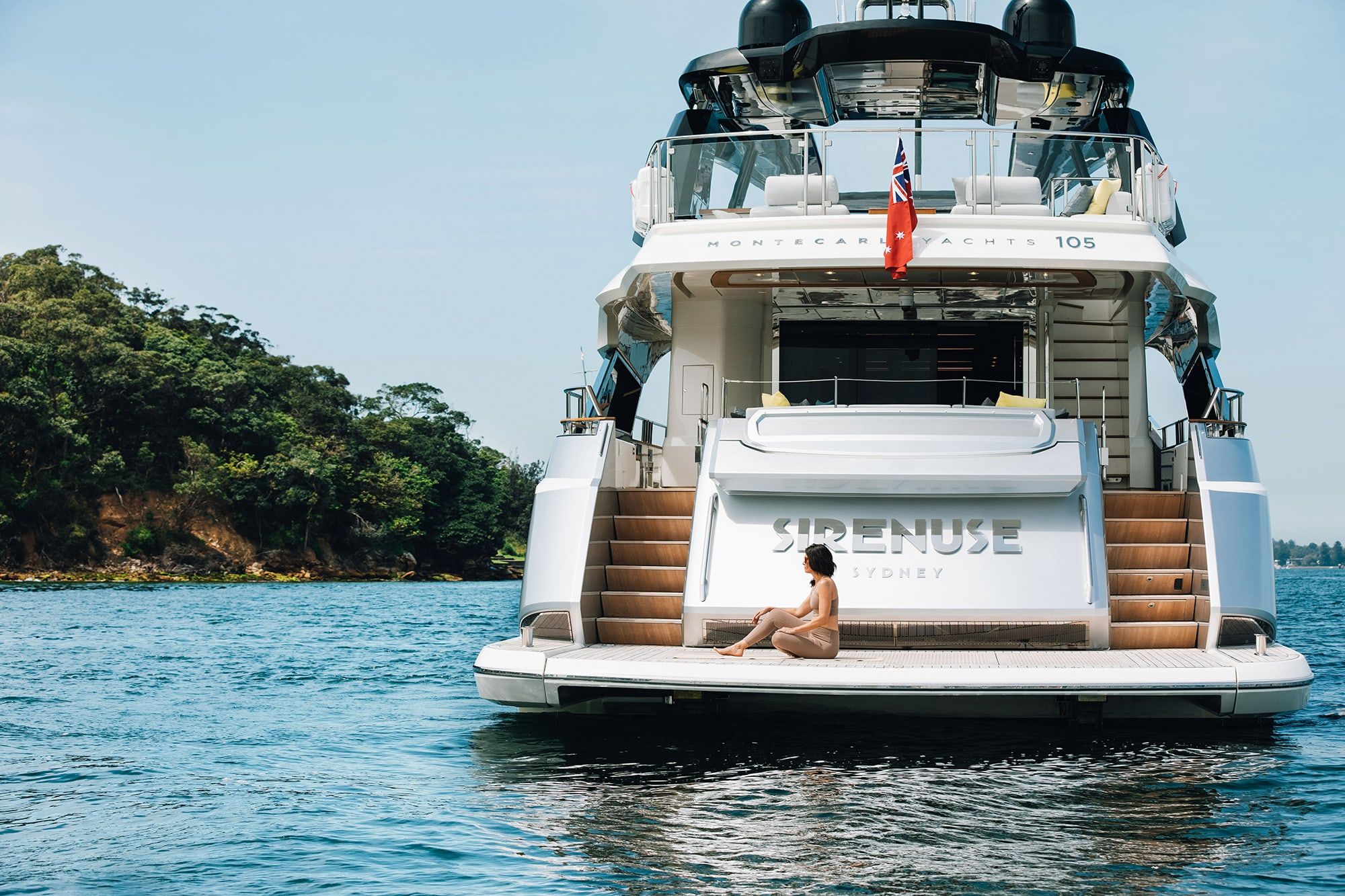 Woman sitting on the swim platform of the motor yacht Sirenuse, anchored near a wooded shoreline
