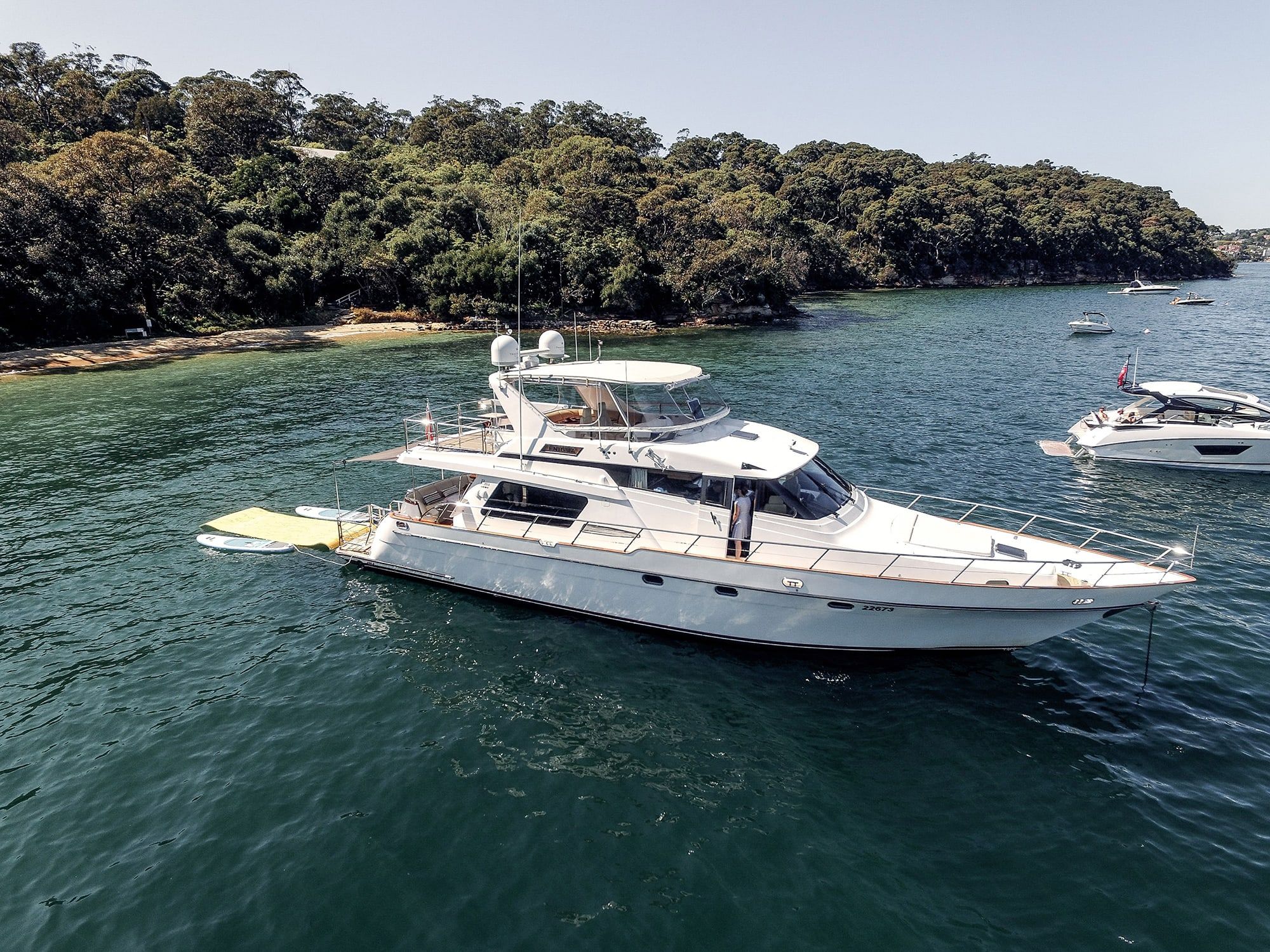 Aerial view of the motor yacht Enigma anchored in green water near a bushland shore, with one person on deck.