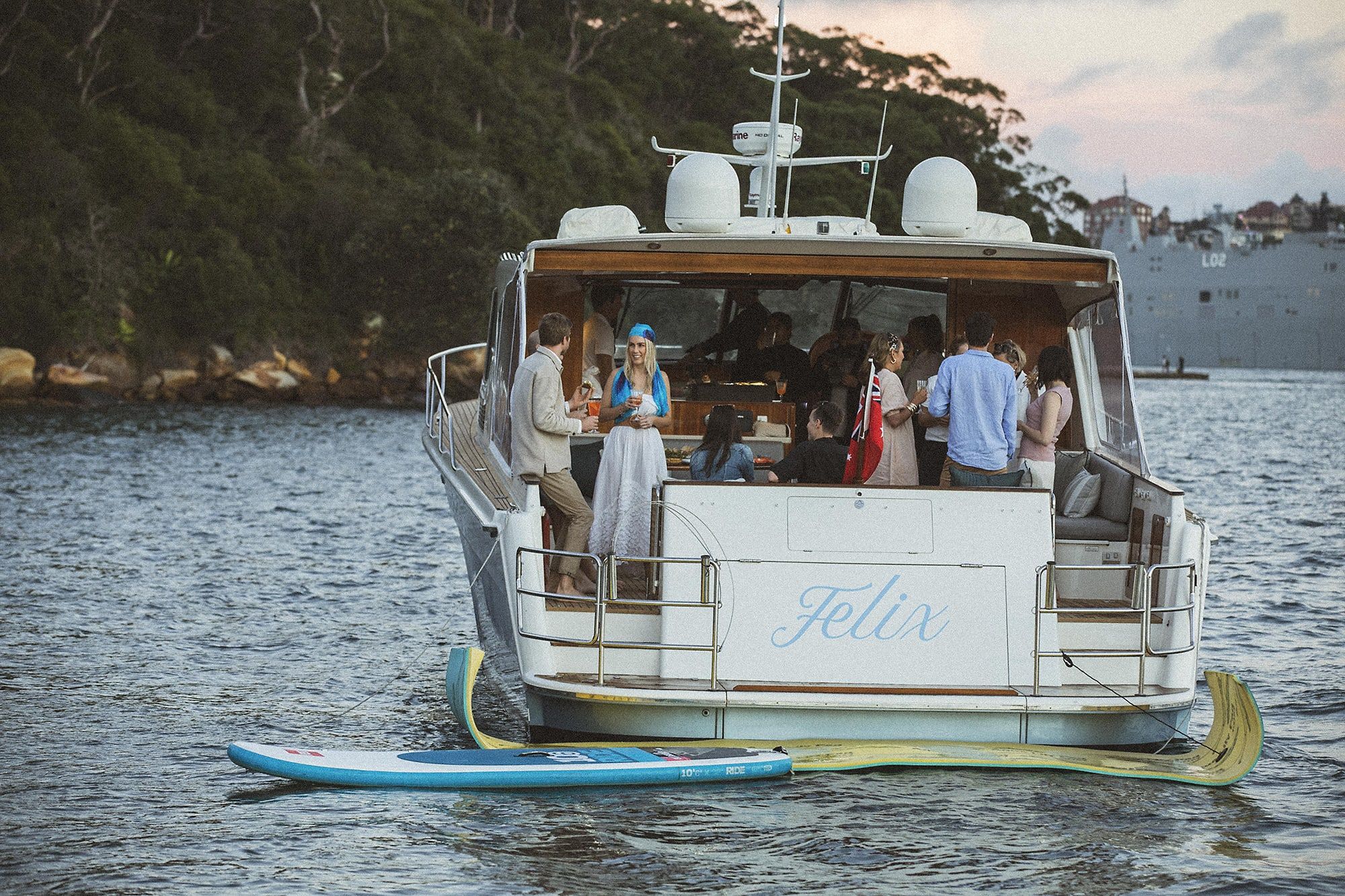 Guests standing with drinks on the aft deck of the motor yacht Felix anchored in a calm bay at dusk.