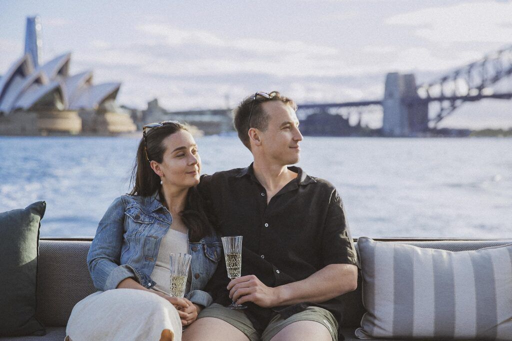 Winter boat party sydney: Couple sitting on a boat's cushioned bench with sparkling wine, harbour and bridge behind them