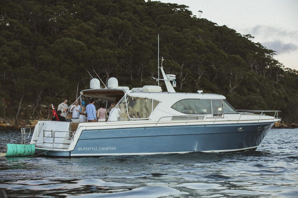 Corporate events sydney: Blue and white motor yacht Felix moored near a bushland shore with a group of people on the aft deck