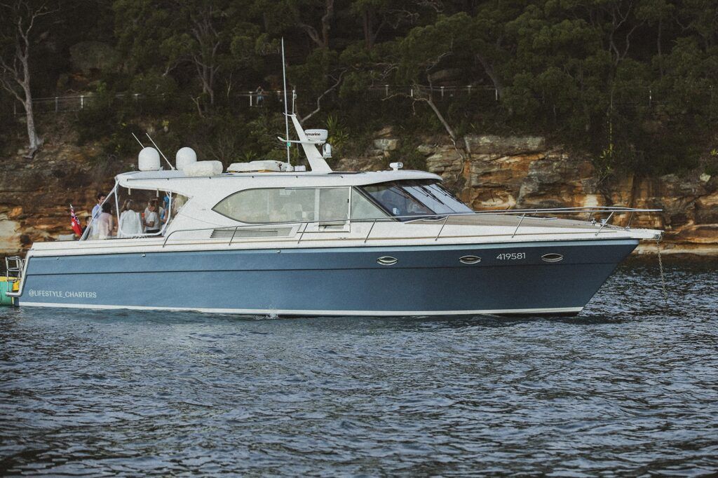 Boat hire manly: Navy-hulled motor yacht Felix anchored side-on in calm water below a sandstone cliff and bushland