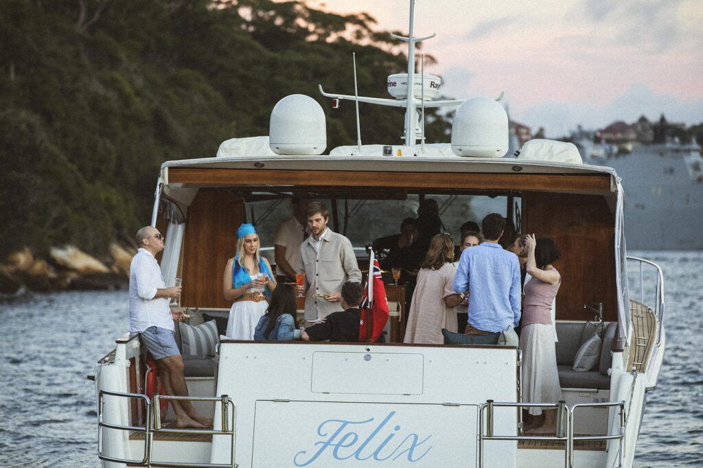 A vibrant group of people enjoy a leisurely gathering on a yacht named ‘Felix’ as it cruises along a scenic waterway. The gue