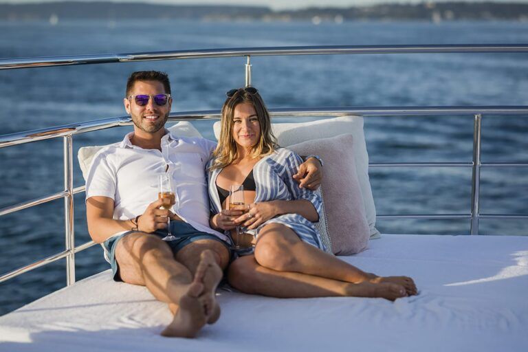 Couple reclining on a white sun lounge on a boat's rear deck holding champagne flutes over the water