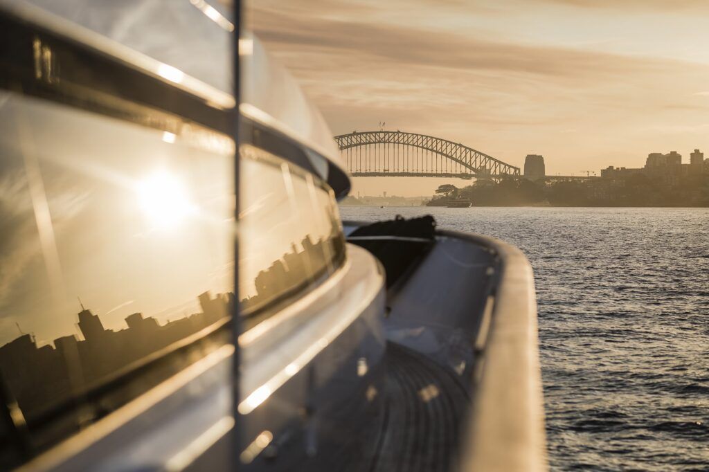 Hiring a boat sydney: View along a motor yacht's side deck at sunset towards a steel arch bridge and city skyline across the