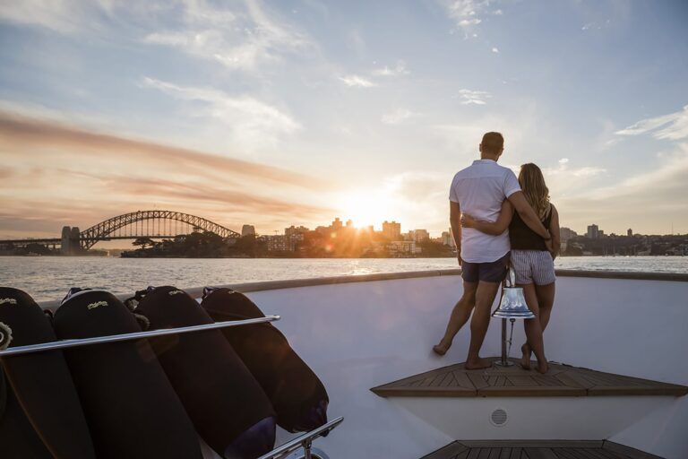 Couple stand arm in arm on the bow of the motor yacht Mohasuwei watching a sunset over a harbour bridge and city skyline.
