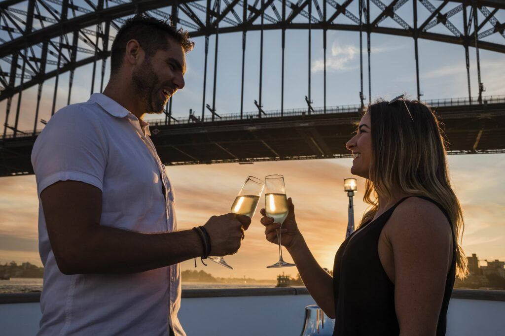 Rose bay wharf: A man and woman smiling as they clink champagne glasses on a boat deck beneath a steel arch bridge at sunset