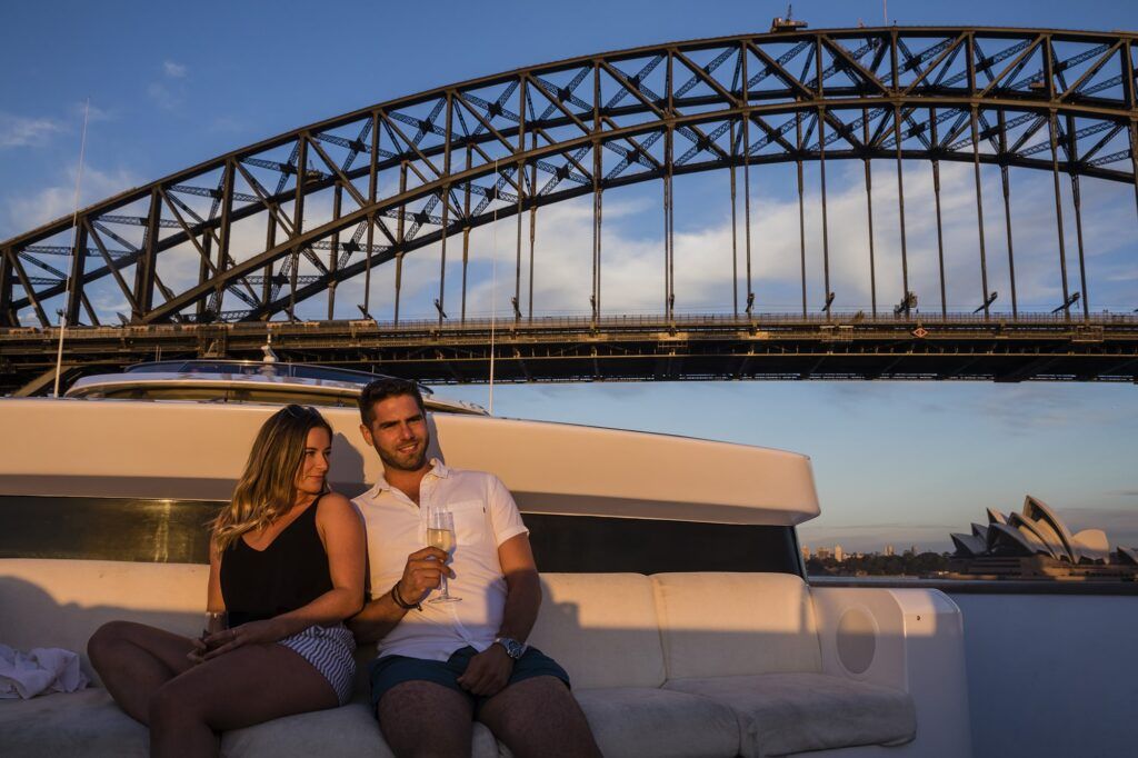 Boat hire sydney: Couple sitting with sparkling wine on a boat's rear lounge, steel arch bridge and opera house behind