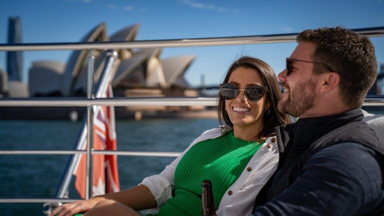 Two people in sunglasses smiling on a boat deck, with the Sydney Opera House and harbour behind them.
