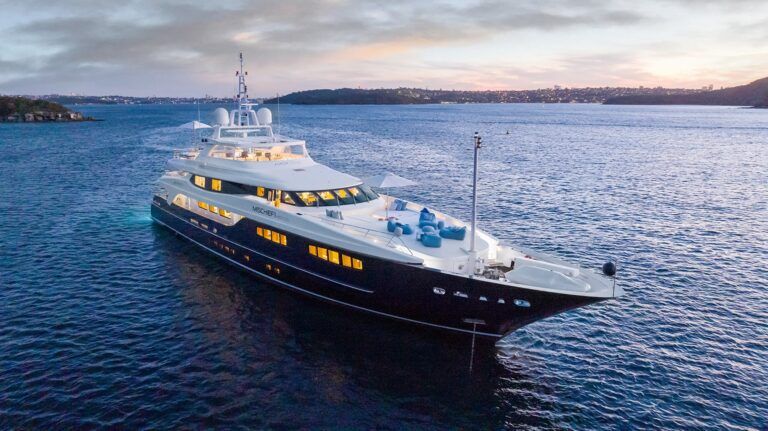 Superyacht Mischief anchored on calm harbour water at dusk with lit interior windows.