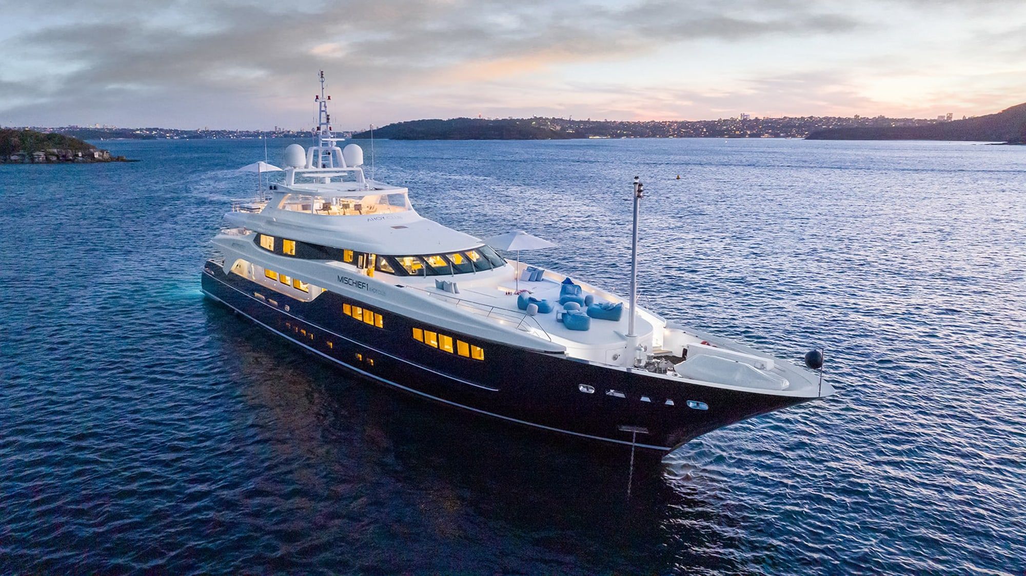 Superyacht Mischief anchored on calm harbour water at dusk with lit interior windows.