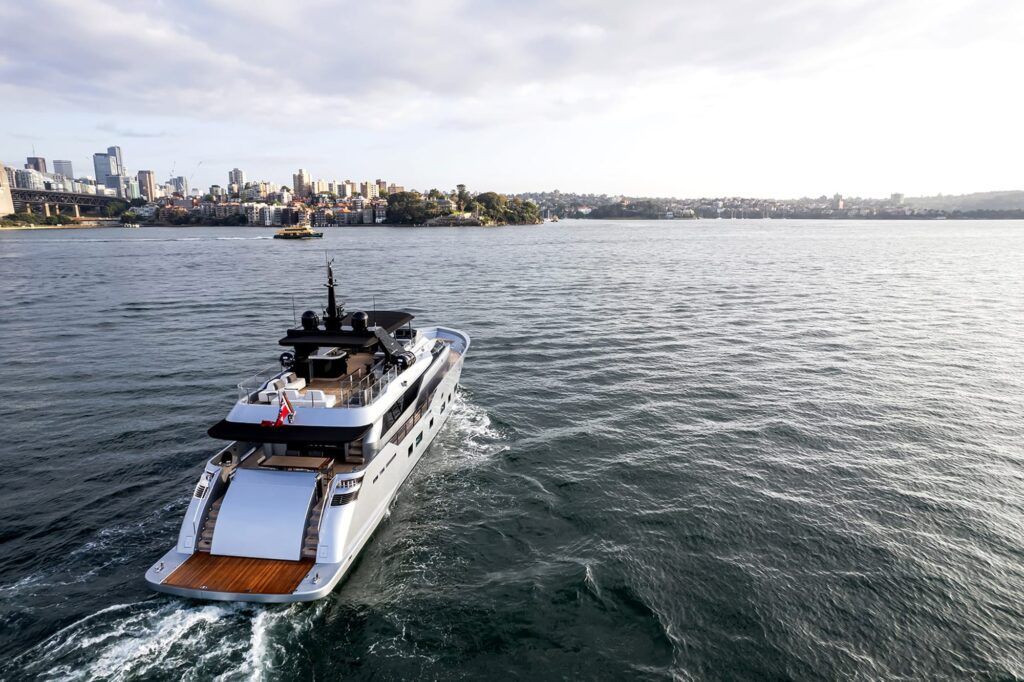 Harbour beach: Aerial view from astern of a white motor yacht underway on a harbour, with a city skyline in the distance