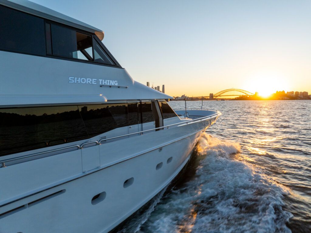 Redleaf beach sydney: White motor yacht named Shore Thing cruising on harbour water at sunset with an arch bridge behind