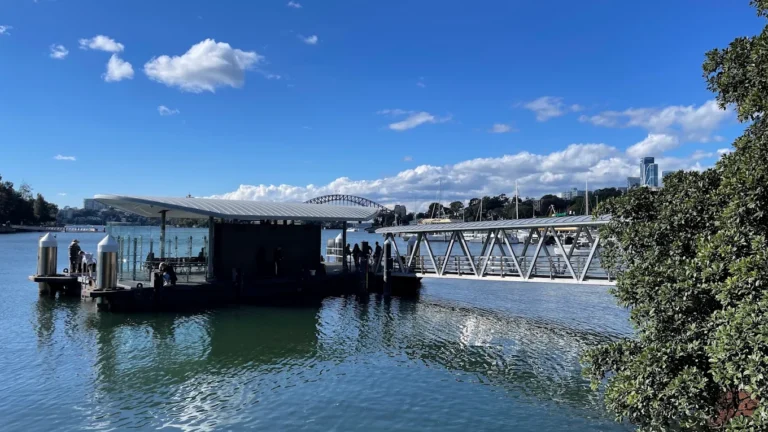 Ferry wharf with a curved metal roof on calm harbour water, with a steel arch bridge in the distance.